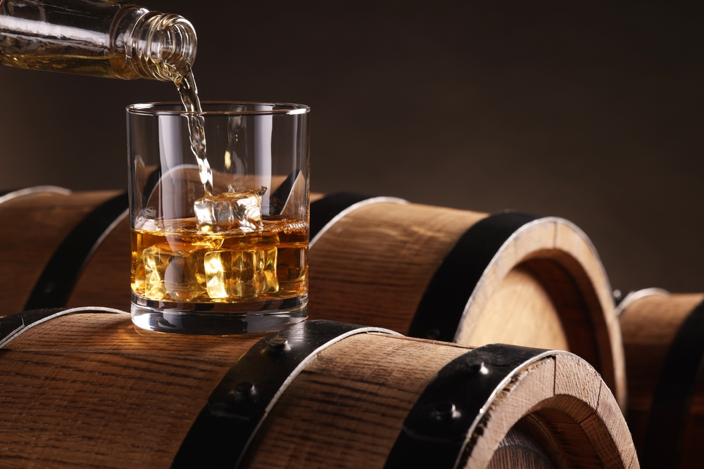 Pouring,Whiskey,From,Bottle,Into,Glass,On,Wooden,Barrel,Against